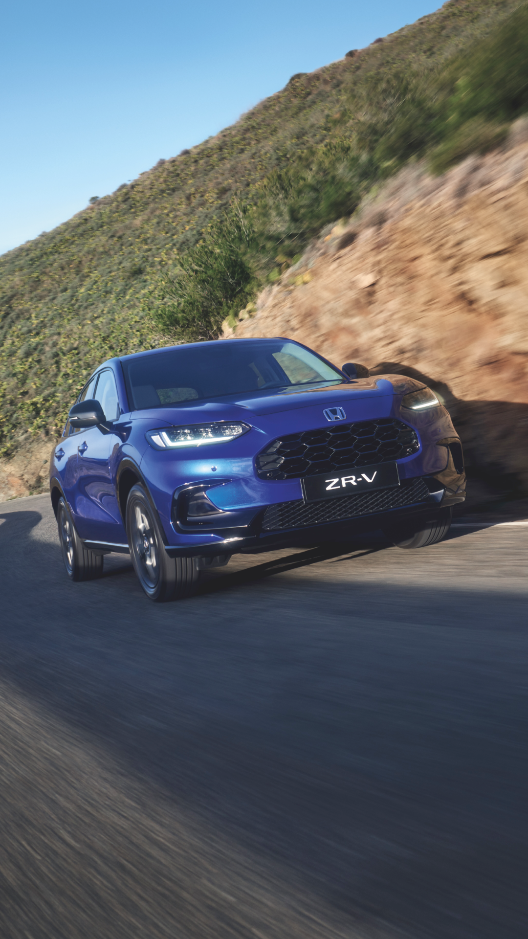 A blue Honda ZR-V SUV driving on a winding mountain road. The car is in motion, with a blurred rocky hillside in the background under a clear blue sky.